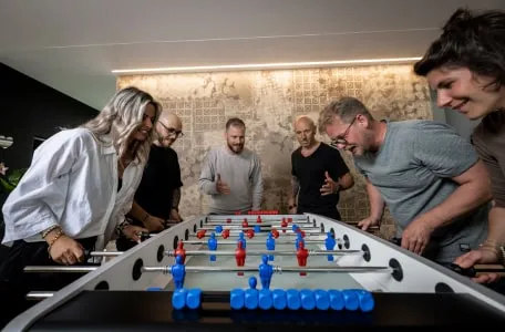 Eine Gruppe von Personen spielt Tischfußball in einem Büro, umgeben von modernen Arbeitsplätzen und entspannter Atmosphäre.